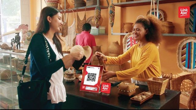 A shopper paying at a craft shop counter by scanning a QR code stand while the shopkeeper hands over a purchase